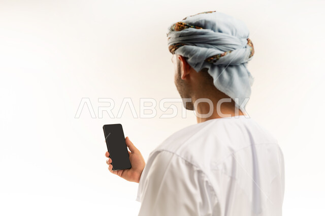 الدردشة مع الأهل والأصدقاء، استخدام جهاز تقني حديث، بورتريه مقرب من الخلف لشاب عربي خليجي عماني يرتدي الدشداشة والعمامة يمسك بيده الهاتف المحمول، دمج التقنية والتكنولوجيا بالحياة اليومية، تصفح مواقع التواصل الاجتماعي، خلفية بيضاء
