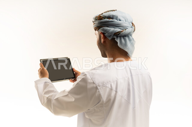 Using modern technology and techniques, browsing social networking sites on a tablet, chatting with family and friends, close-up portrait from the back of an Arab Gulf Omani man wearing a dishdasha and turban holding a tablet, white background