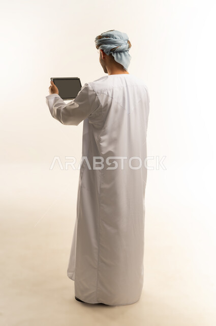 استعمال التكنولوجيا والتقنية الحديثة, تصفح مواقع التواصل الاجتماعي عبر الجهاز اللوحي، المحادثة مع الأهل والأصدقاء، بورتريه من الخلف لرجل عربي خليجي عماني يرتدي الدشداشة والعمامة يحمل بيده جهاز التابلت، الطول الكامل للجسم، خلفية بيضاء