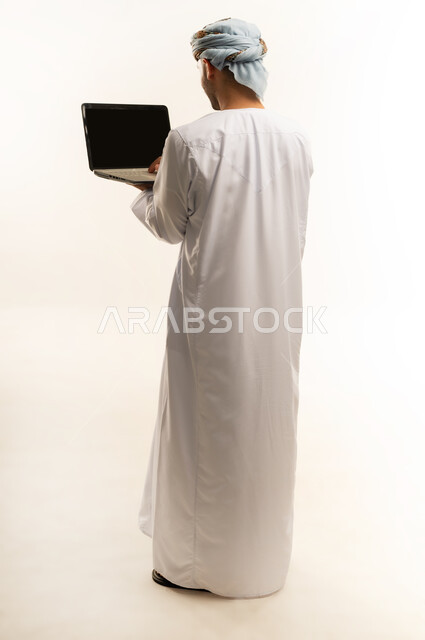 Using modern technology, the concept of diligence and completing required tasks, a portrait from the back of an Arab Gulf Omani young man wearing a dishdasha and turban, standing and holding a laptop in his hand, browsing social networking sites, integrating technology into daily life, a full-length body image, white background
