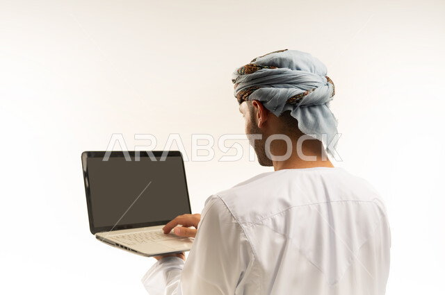 Using modern technology, the concept of diligence and completing required tasks, a close-up portrait from the back of an Arab Gulf Omani young man wearing a dishdasha and turban, standing and holding a laptop in his hand, browsing social media sites, integrating technology into daily life, white background