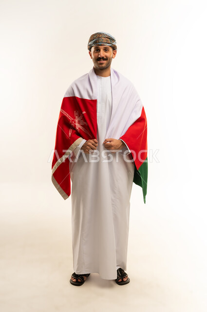 إحياء ذكرى تأسيس الدولة 1775 م،  ثقافات و رموز وطنية, حب الوطن والانتماء إليه، العلم الوطني للبلاد، بورتريه لرجل عربي خليجي عماني يرتدي الدشداشة و العمامة التقليدية يحتفل بذكرى اليوم الوطني العماني 18 نوفمبر، الجسم بالطول الكامل، خلفية بيضاء