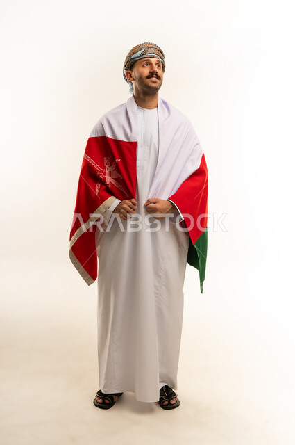 ثقافات و رموز وطنية, إحياء ذكرى تأسيس الدولة 1775 م، حب الوطن والانتماء إليه، العلم الوطني للبلاد، بورتريه لرجل عربي خليجي عماني يرتدي الدشداشة و العمامة التقليدية يحتفل بذكرى اليوم الوطني العماني 18 نوفمبر، الجسم بالطول الكامل، خلفية بيضاء
