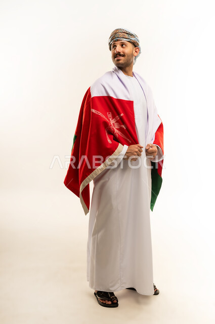 ثقافات و رموز وطنية, إحياء ذكرى تأسيس الدولة 1775 م، حب الوطن والانتماء إليه، العلم الوطني للبلاد، بورتريه لرجل عربي خليجي عماني يرتدي الدشداشة و العمامة التقليدية يحتفل بذكرى اليوم الوطني العماني 18 نوفمبر، الجسم بالطول الكامل، خلفية بيضاء