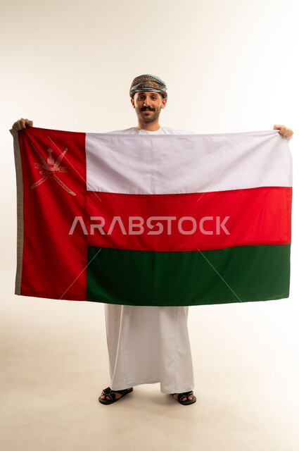 إحياء ذكرى تأسيس الدولة 1775 م،  ثقافات و رموز وطنية, حب الوطن والانتماء إليه، العلم الوطني للبلاد، بورتريه لرجل عربي خليجي عماني يرتدي الدشداشة و العمامة التقليدية يحتفل بذكرى اليوم الوطني العماني 18 نوفمبر، الجسم بالطول الكامل، خلفية بيضاء