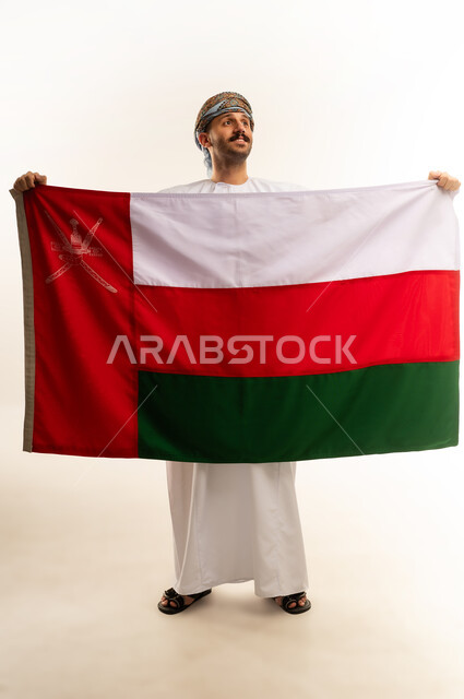 ثقافات و رموز وطنية, إحياء ذكرى تأسيس الدولة 1775 م، حب الوطن والانتماء إليه، العلم الوطني للبلاد، بورتريه لرجل عربي خليجي عماني يرتدي الدشداشة و العمامة التقليدية يحتفل بذكرى اليوم الوطني العماني 18 نوفمبر، الجسم بالطول الكامل، خلفية بيضاء
