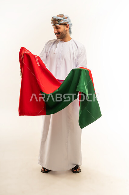 إحياء ذكرى تأسيس الدولة 1775 م،  ثقافات و رموز وطنية, حب الوطن والانتماء إليه، العلم الوطني للبلاد، بورتريه لرجل عربي خليجي عماني يرتدي الدشداشة و العمامة التقليدية يحتفل بذكرى اليوم الوطني العماني 18 نوفمبر، الجسم بالطول الكامل، خلفية بيضاء