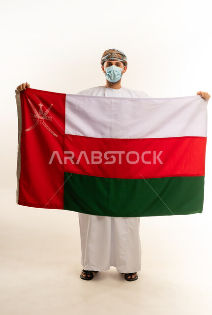 إحياء ذكرى تأسيس الدولة 1775 م،  ثقافات و رموز وطنية, حب الوطن والانتماء إليه، العلم الوطني للبلاد، بورتريه لرجل عربي خليجي عماني يرتدي الدشداشة و العمامة التقليدية يحتفل بذكرى اليوم الوطني العماني 18 نوفمبر، الجسم بالطول الكامل، خلفية بيضاء