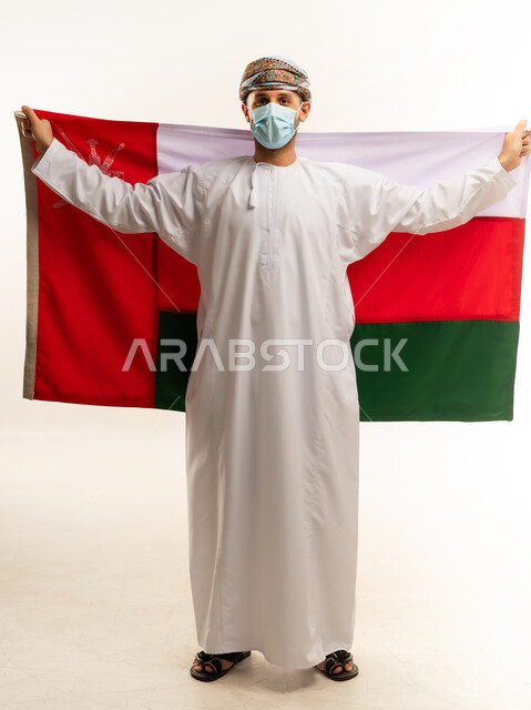 ثقافات و رموز وطنية, إحياء ذكرى تأسيس الدولة 1775 م، حب الوطن والانتماء إليه، العلم الوطني للبلاد، بورتريه لرجل عربي خليجي عماني يرتدي الدشداشة و العمامة التقليدية يحتفل بذكرى اليوم الوطني العماني 18 نوفمبر، الجسم بالطول الكامل، خلفية بيضاء
