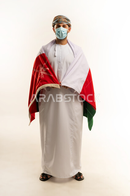 ثقافات و رموز وطنية, إحياء ذكرى تأسيس الدولة 1775 م، حب الوطن والانتماء إليه، العلم الوطني للبلاد، بورتريه لرجل عربي خليجي عماني يرتدي الدشداشة و العمامة التقليدية يحتفل بذكرى اليوم الوطني العماني 18 نوفمبر، الجسم بالطول الكامل، خلفية بيضاء