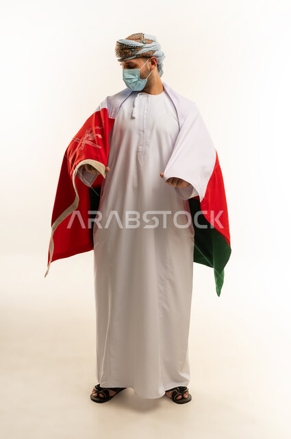 إحياء ذكرى تأسيس الدولة 1775 م،  ثقافات و رموز وطنية, حب الوطن والانتماء إليه، العلم الوطني للبلاد، بورتريه لرجل عربي خليجي عماني يرتدي الدشداشة و العمامة التقليدية يحتفل بذكرى اليوم الوطني العماني 18 نوفمبر، الجسم بالطول الكامل، خلفية بيضاء