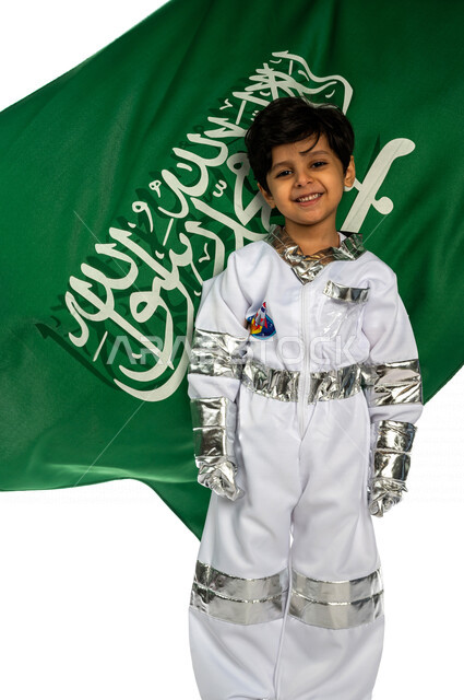 بورتريه لفتى عربي خليجي سعودي مبتسم ، يرتدي زي رائد الفضاء ، ينظر إلى الكاميرا ، خلفه علم المملكة العربية السعودية ، ذكرى اليوم الوطني السعودي ،الاحتفال بالعيد الوطني السعودي ، يحلم بأن يصبح رائد فضاء ، حلم الوصول للفضاء والقمر ، خلفية بيضاء