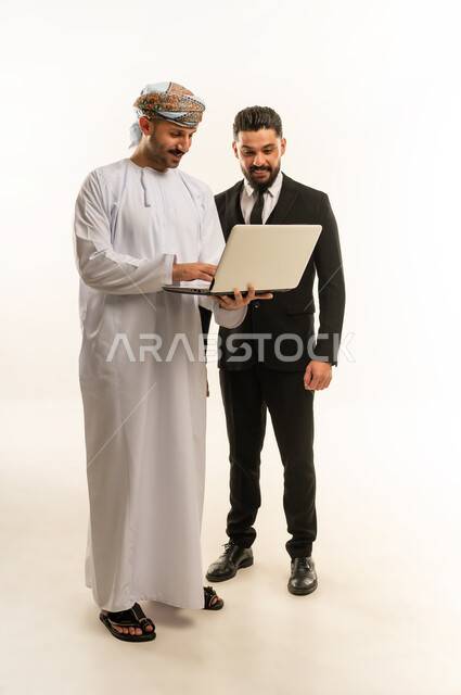 استخدام التقنية في العمل، استضافة رجال الاعمال الاجانب في سلطنة عمان, تبادل الخبرات والمعرفة بين البلاد, بورتريه لرجل عربي خليجي عماني يرتدي العمامة والدشداشة يقف بجانب شاب اجبني يرتدي البدلة الرسمية يقومان بالعمل على الحاسوب المحمول، الجسم بالطول الكامل، خلفية بيضاء