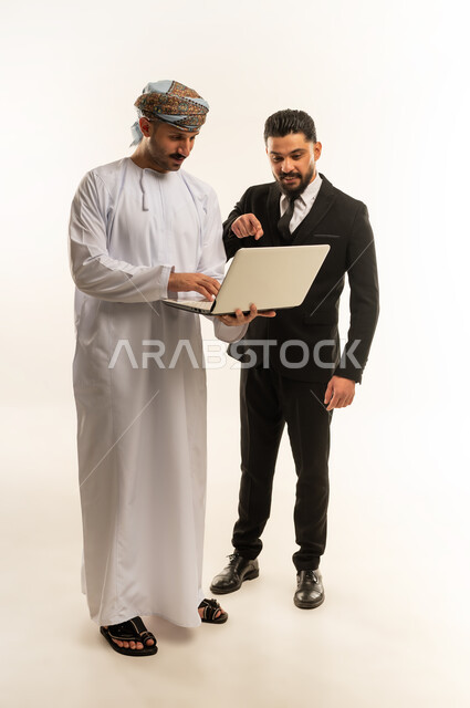 Using technology at work, hosting foreign businessmen in the Sultanate of Oman, exchanging experiences and knowledge between countries, portrait of an Arab Gulf Omani man wearing a turban and dishdasha standing next to a young man wearing a formal suit, both working on a laptop, full body, white background