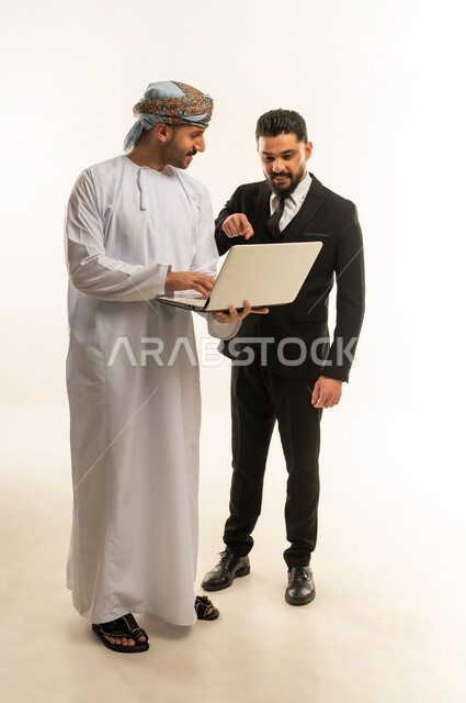 Using technology at work, hosting foreign businessmen in the Sultanate of Oman, exchanging experiences and knowledge between countries, portrait of an Arab Gulf Omani man wearing a turban and dishdasha standing next to a young man wearing a formal suit, both working on a laptop, full body, white background