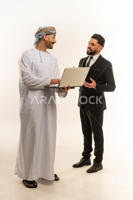 استخدام التقنية في العمل، استضافة رجال الاعمال الاجانب في سلطنة عمان, تبادل الخبرات والمعرفة بين البلاد, بورتريه لرجل عربي خليجي عماني يرتدي العمامة والدشداشة يقف بجانب شاب اجبني يرتدي البدلة الرسمية يقومان بالعمل على الحاسوب المحمول، الجسم بالطول الكامل، خلفية بيضاء
