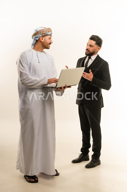 استخدام التقنية في العمل، استضافة رجال الاعمال الاجانب في سلطنة عمان, تبادل الخبرات والمعرفة بين البلاد, بورتريه لرجل عربي خليجي عماني يرتدي العمامة والدشداشة يقف بجانب شاب اجبني يرتدي البدلة الرسمية يقومان بالعمل على الحاسوب المحمول، الجسم بالطول الكامل، خلفية بيضاء