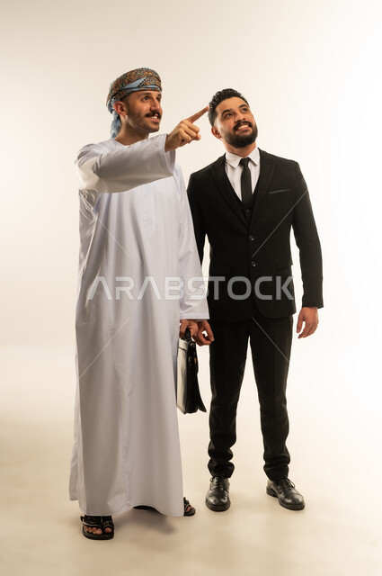استضافة رجال الاعمال الاجانب في سلطنة عمان, تبادل الخبرات والمعرفة بين البلاد, بورتريه لرجل عربي خليجي عماني يرتدي الدشداشة والعمامة يقف بجانب سائح اجبني يرتدي البدلة الرسمية ينظران الى شيء ما بتعابير السرور, صورة لكامل الجسم، خلفية بيضاء
