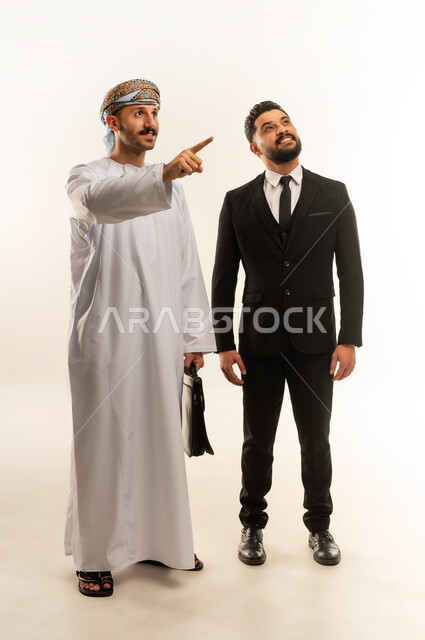 استضافة رجال الاعمال الاجانب في سلطنة عمان, تبادل الخبرات والمعرفة بين البلاد, بورتريه لرجل عربي خليجي عماني يرتدي الدشداشة والعمامة يقف بجانب سائح اجبني يرتدي البدلة الرسمية ينظران الى شيء ما بتعابير السرور, صورة لكامل الجسم، خلفية بيضاء