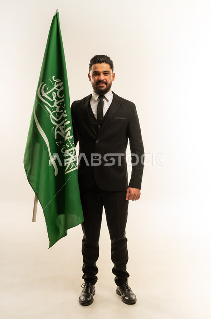 ذكرى يوم العلم 11 مارس، التعرف على ثقافات وتاريخ السعودية، بورتريه لرجل اعمال اجنبي مبتسم يرتدي البدلة الرسمية يحمل العلم للاحتفال باليوم بالوطني السعودي 23 سبتمبر، الوقوف باستقامة وثبات بتعابير الفخر والاعتزاز، النظر إلى الكاميرا بايماءات السرور، خلفية بيضاء