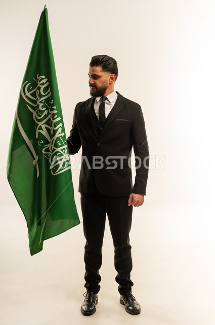 التعرف على ثقافات وتاريخ السعودية، ذكرى يوم العلم 11 مارس، بورتريه لرجل اعمال اجنبي مبتسم يرتدي البدلة الرسمية يحمل العلم للاحتفال باليوم بالوطني السعودي 23 سبتمبر، الوقوف باستقامة وثبات بتعابير الفخر والاعتزاز، النظر إلى الكاميرا بايماءات السرور، خلفية بيضاء