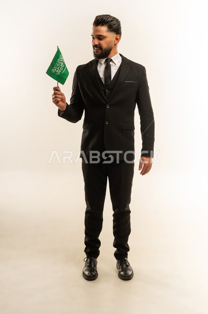 التعرف على ثقافات وتاريخ السعودية، ذكرى يوم العلم 11 مارس، بورتريه لرجل اعمال اجنبي مبتسم يرتدي البدلة الرسمية يحمل العلم للاحتفال باليوم بالوطني السعودي 23 سبتمبر، الوقوف باستقامة وثبات بتعابير الفخر والاعتزاز، النظر إلى الكاميرا بايماءات السرور، خلفية بيضاء