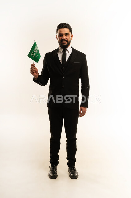 ذكرى يوم العلم 11 مارس، التعرف على ثقافات وتاريخ السعودية، بورتريه لرجل اعمال اجنبي مبتسم يرتدي البدلة الرسمية يحمل العلم للاحتفال باليوم بالوطني السعودي 23 سبتمبر، الوقوف باستقامة وثبات بتعابير الفخر والاعتزاز، النظر إلى الكاميرا بايماءات السرور، خلفية بيضاء