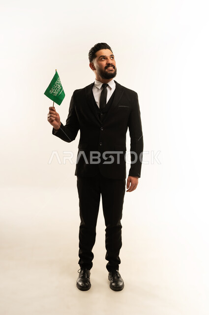 Learning about Saudi culture and history, Flag Day commemoration, March 11, portrait of a smiling foreign businessman wearing a formal suit holding a flag to celebrate Saudi National Day, September 23, standing straight and firm with expressions of pride and honor, looking at the camera with gestures of pleasure, white background