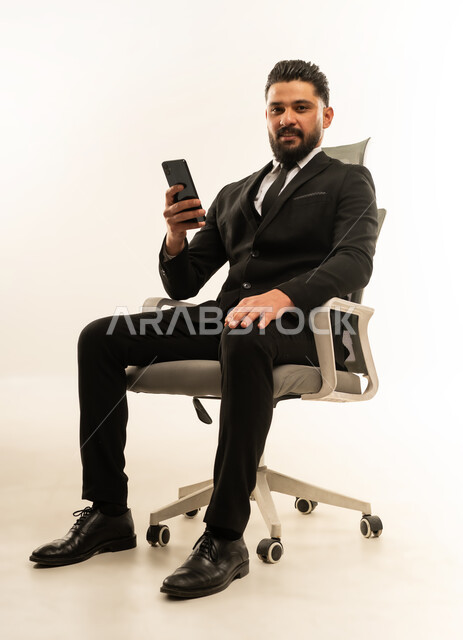Integrating technology into practical and daily life, communicating with family and friends, gestures of happiness and pleasure, portrait of a smiling foreign businessman wearing a formal suit sitting on a chair holding a mobile phone, using modern technologies and devices, white background