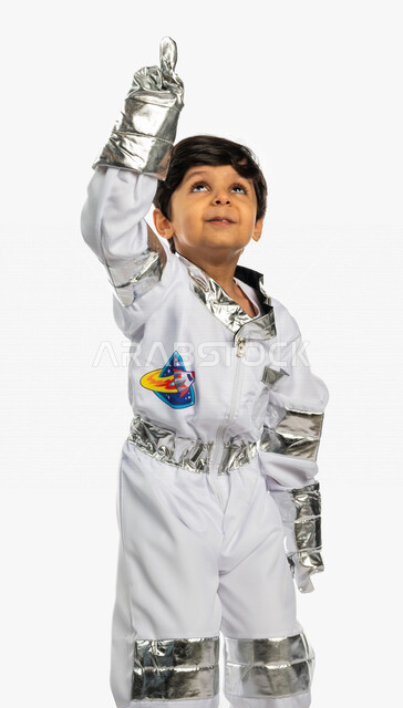 Portrait of a smiling Saudi Arabian Gulf Arab boy, wearing an astronaut costume, raising his hand up, dreaming of becoming an astronaut, pointing his index finger up with different facial and hands gestures indicating happiness, dream of reaching space an