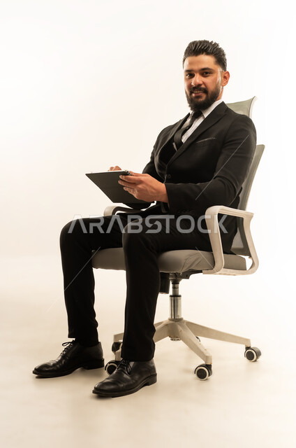 Holding a business meeting, integrating modern devices and technology into work life, browsing the internet and completing tasks remotely, following social media news, portrait of a foreign businessman wearing a formal suit sitting on a chair using a tablet, white background