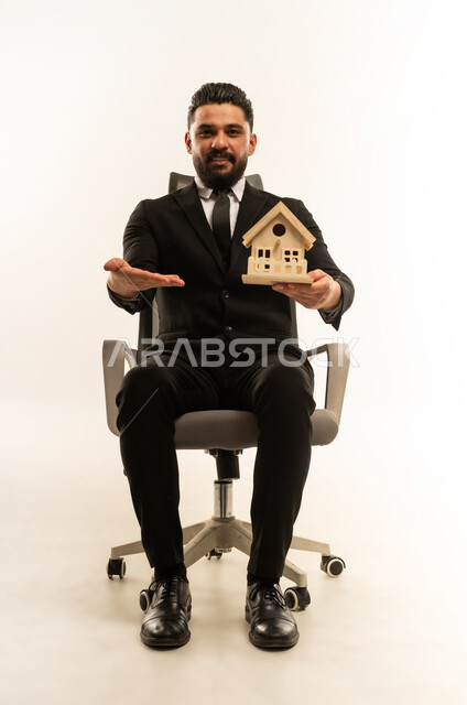 إيماءات السرور والسعادة، السعي لامتلاك بيت جديد، بيع وشراء العقارات، بورتريه لشاب عربي اجنبي يرتدي البدلة الرسمية  يجلس على كنبة مريحة ويحمل بيديه مجسم خشبي صغير لمنزل يشير اليه بيده، خلفية بيضاء