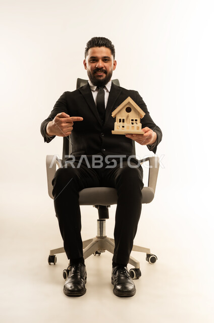 إيماءات السرور والسعادة، السعي لامتلاك بيت جديد، بيع وشراء العقارات، بورتريه لشاب عربي اجنبي يرتدي البدلة الرسمية  يجلس على كنبة مريحة ويحمل بيديه مجسم خشبي صغير لمنزل يشير اليه بيده، خلفية بيضاء