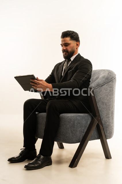 Holding a business meeting, integrating modern devices and technology into work life, browsing the internet and completing tasks remotely, following social media news, portrait of a foreign businessman wearing a formal suit sitting on a chair using a tablet, white background