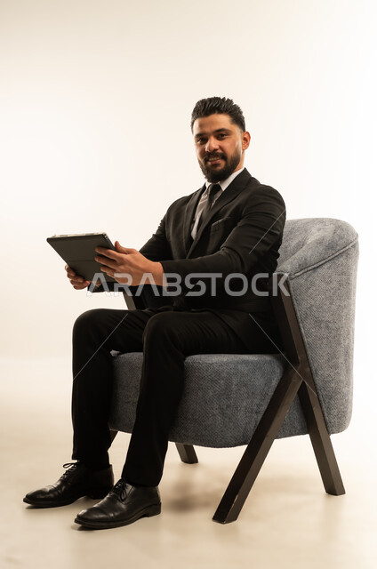 Holding a business meeting, integrating modern devices and technology into work life, browsing the internet and completing tasks remotely, following social media news, portrait of a foreign businessman wearing a formal suit sitting on a chair using a tablet, white background