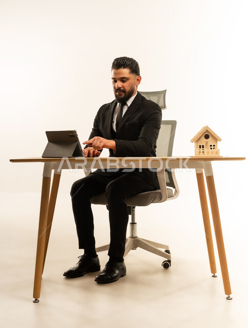 بيع وشراء العقارات عبر الانترنت، إيماءات السرور والسعادة، السعي لامتلاك بيت جديد، بورتريه لشاب عربي اجنبي يرتدي البدلة الرسمية  يجلس على مكتبه يعمل على جهاز التابلت اللوحي، مجسم خشبي صغير لمنزل، خلفية بيضاء