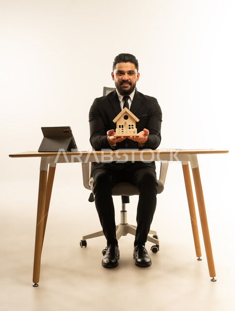 بيع وشراء العقارات عبر الانترنت، إيماءات السرور والسعادة، السعي لامتلاك بيت جديد، بورتريه لشاب عربي اجنبي يرتدي البدلة الرسمية  يجلس على مكتبه يعمل على جهاز التابلت اللوحي، مجسم خشبي صغير لمنزل، خلفية بيضاء
