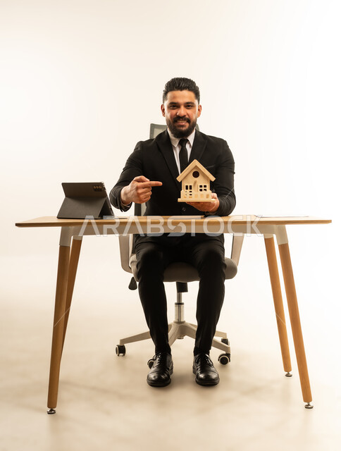 بيع وشراء العقارات عبر الانترنت، إيماءات السرور والسعادة، السعي لامتلاك بيت جديد، بورتريه لشاب عربي اجنبي يرتدي البدلة الرسمية  يجلس على مكتبه يعمل على جهاز التابلت اللوحي، مجسم خشبي صغير لمنزل، خلفية بيضاء