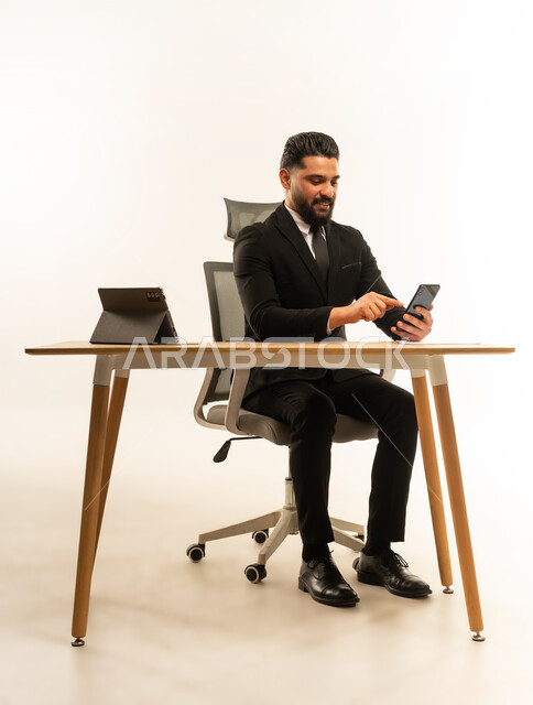 دمج التكنولوجيا في الحياة العملية واليومية، التواصل مع الاهل والاصدقاء، ايماءات السعادة والسرور، بورتريه لرجل اعمال اجنبي مبتسم يرتدي البدلة الرسمية يجلس على مكتبه امام جهاز التايلت يمسك بيده الهاتف المحمول، استخدام التقنيات والاجهزة الحديثة، خلفية بيضاء