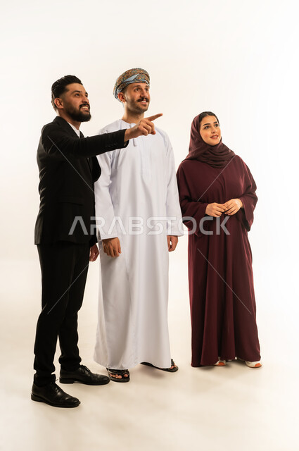 الاشارة الى شيء ما بتعابير السرور والسعادة، بورتريه لرجل عربي خليجي عماني يرتدي الدشداشة والعمامة يقف بجانب إمرأة وشاب اجنبي يرتدي البدلة الرسمية، الوقوف باستقامة والنظر لمكان ما، الجسم بالطول الكامل، خلفية بيضاء