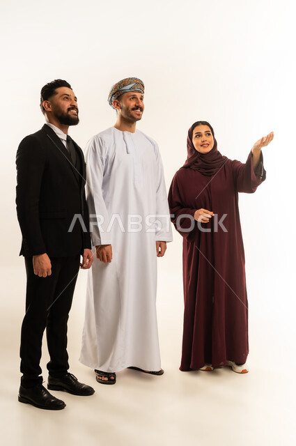 الاشارة الى شيء ما بتعابير السرور والسعادة، بورتريه لرجل عربي خليجي عماني يرتدي الدشداشة والعمامة يقف بجانب إمرأة وشاب اجنبي يرتدي البدلة الرسمية، الوقوف باستقامة والنظر لمكان ما، الجسم بالطول الكامل، خلفية بيضاء