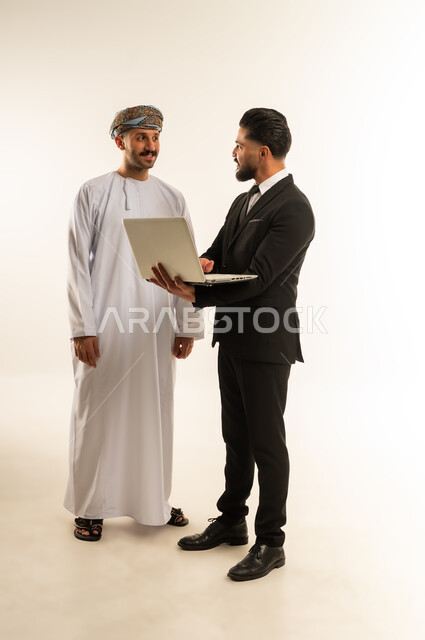 استضافة رجال الاعمال الاجانب في سلطنة عمان، تبادل الخبرات والمعرفة بين البلاد, بورتريه لرجل عربي خليجي عماني يرتدي الدشداشة والعمامة يقف بجانب شاب اجبني يرتدي البدلة الرسمية يقومان بالعمل على الحاسب المحمول، صورة لكامل الجسم، خلفية بيضاء