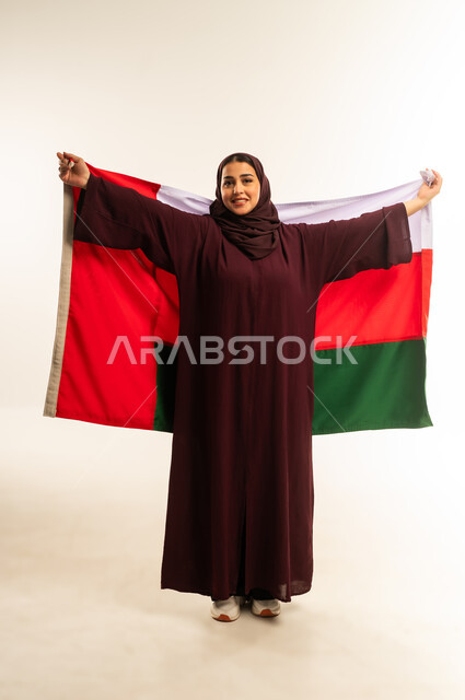 حب الوطن والانتماء إليه، إحياء ذكرى تأسيس الدولة 1775 م، ثقافات ورموز وطنية, بورتريه لإمرأة عربية خليجية عمانية ترتدي الحجاب والعباءة تمسك  العلم الوطني للبلاد تحتفل بذكرى اليوم الوطني العماني 18 نوفمبر، الجسم بالطول الكامل، خلفية بيضاء