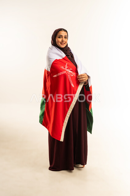 حب الوطن والانتماء إليه، إحياء ذكرى تأسيس الدولة 1775 م، ثقافات ورموز وطنية, بورتريه لإمرأة عربية خليجية عمانية ترتدي الحجاب والعباءة تمسك  العلم الوطني للبلاد تحتفل بذكرى اليوم الوطني العماني 18 نوفمبر، الجسم بالطول الكامل، خلفية بيضاء