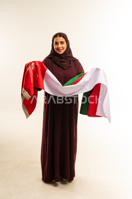 حب الوطن والانتماء إليه، إحياء ذكرى تأسيس الدولة 1775 م، ثقافات ورموز وطنية, بورتريه لإمرأة عربية خليجية عمانية ترتدي الحجاب والعباءة تمسك  العلم الوطني للبلاد تحتفل بذكرى اليوم الوطني العماني 18 نوفمبر، الجسم بالطول الكامل، خلفية بيضاء