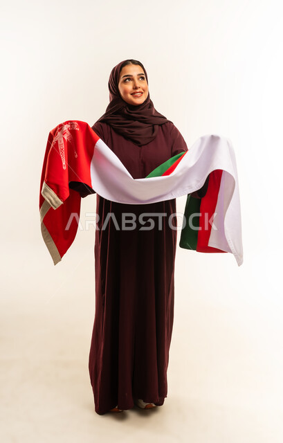 إحياء ذكرى تأسيس الدولة 1775 م، حب الوطن والانتماء إليه، ثقافات ورموز وطنية, بورتريه لإمرأة عربية خليجية عمانية ترتدي الحجاب والعباءة تمسك  العلم الوطني للبلاد تحتفل بذكرى اليوم الوطني العماني 18 نوفمبر، الجسم بالطول الكامل، خلفية بيضاء