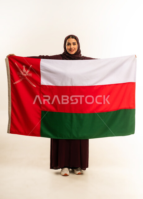 حب الوطن والانتماء إليه، إحياء ذكرى تأسيس الدولة 1775 م، ثقافات ورموز وطنية, بورتريه لإمرأة عربية خليجية عمانية ترتدي الحجاب والعباءة تمسك  العلم الوطني للبلاد تحتفل بذكرى اليوم الوطني العماني 18 نوفمبر، الجسم بالطول الكامل، خلفية بيضاء