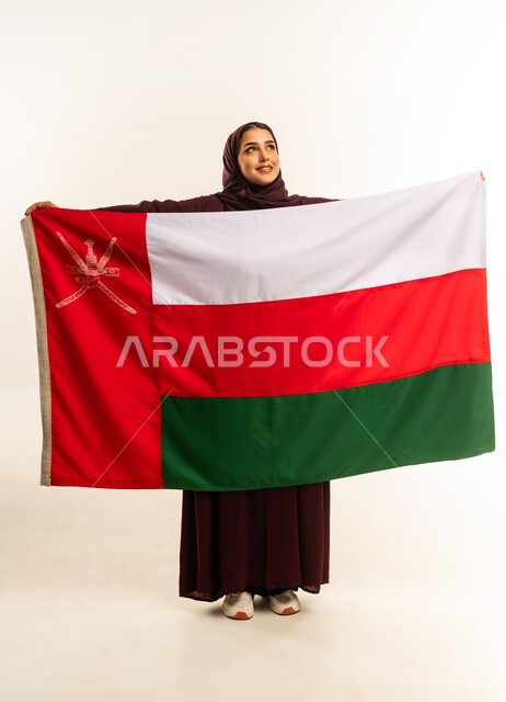إحياء ذكرى تأسيس الدولة 1775 م، حب الوطن والانتماء إليه، ثقافات ورموز وطنية, بورتريه لإمرأة عربية خليجية عمانية ترتدي الحجاب والعباءة تمسك  العلم الوطني للبلاد تحتفل بذكرى اليوم الوطني العماني 18 نوفمبر، الجسم بالطول الكامل، خلفية بيضاء