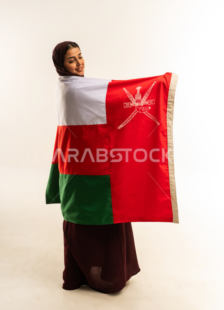 حب الوطن والانتماء إليه، إحياء ذكرى تأسيس الدولة 1775 م، ثقافات ورموز وطنية, بورتريه لإمرأة عربية خليجية عمانية ترتدي الحجاب والعباءة تمسك  العلم الوطني للبلاد تحتفل بذكرى اليوم الوطني العماني 18 نوفمبر، الجسم بالطول الكامل، خلفية بيضاء