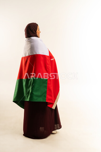 إحياء ذكرى تأسيس الدولة 1775 م، حب الوطن والانتماء إليه، ثقافات ورموز وطنية, بورتريه من الخلف لإمرأة عربية خليجية عمانية ترتدي الحجاب والعباءة والعلم الوطني للبلاد تحتفل بذكرى اليوم الوطني العماني 18 نوفمبر، الجسم بالطول الكامل، خلفية بيضاء